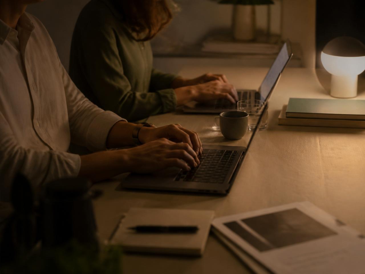 Two people working late at night on laptops, warm lamp light.